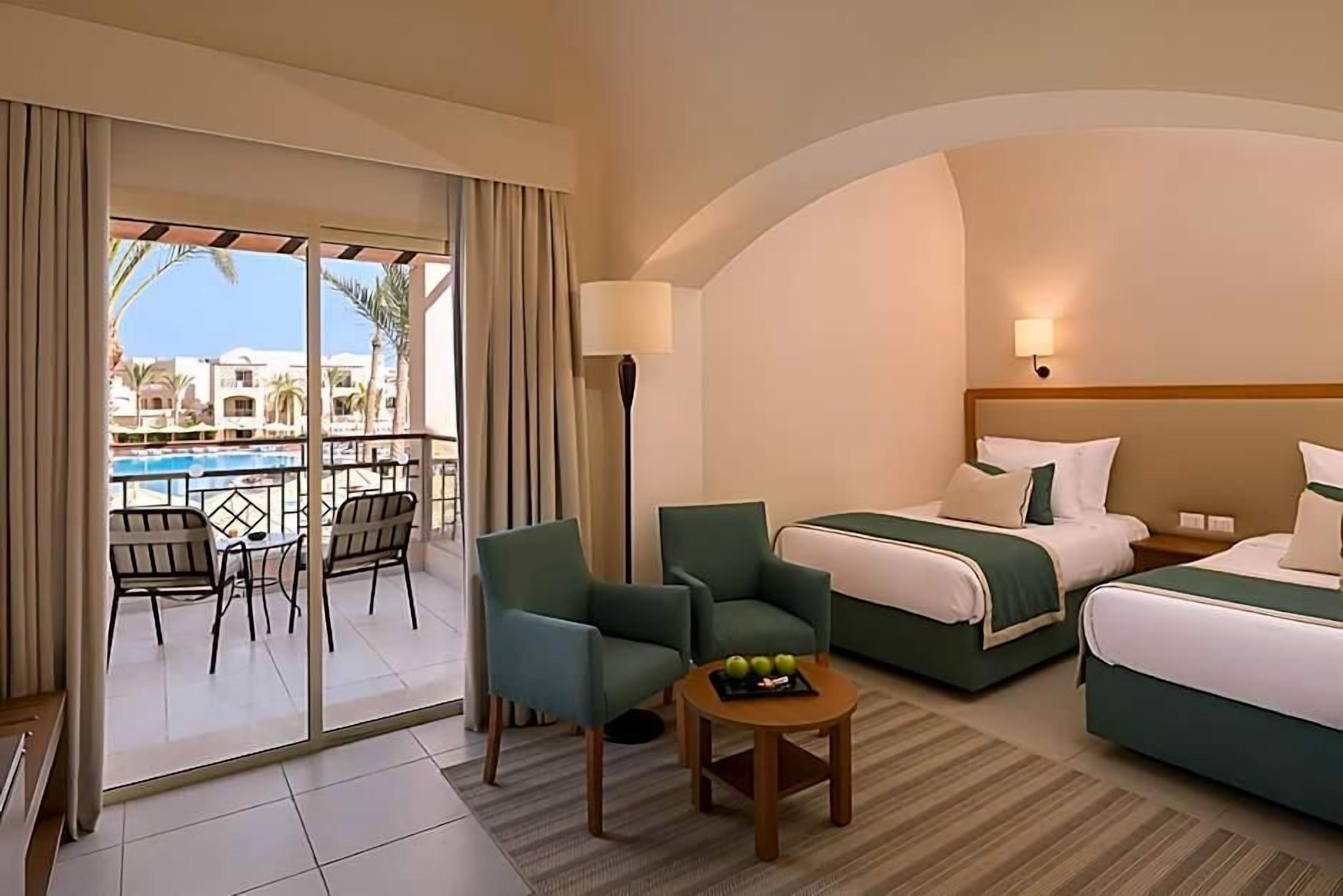 A hotel room with a balcony, two beds, a table with fruit, and a view of buildings.