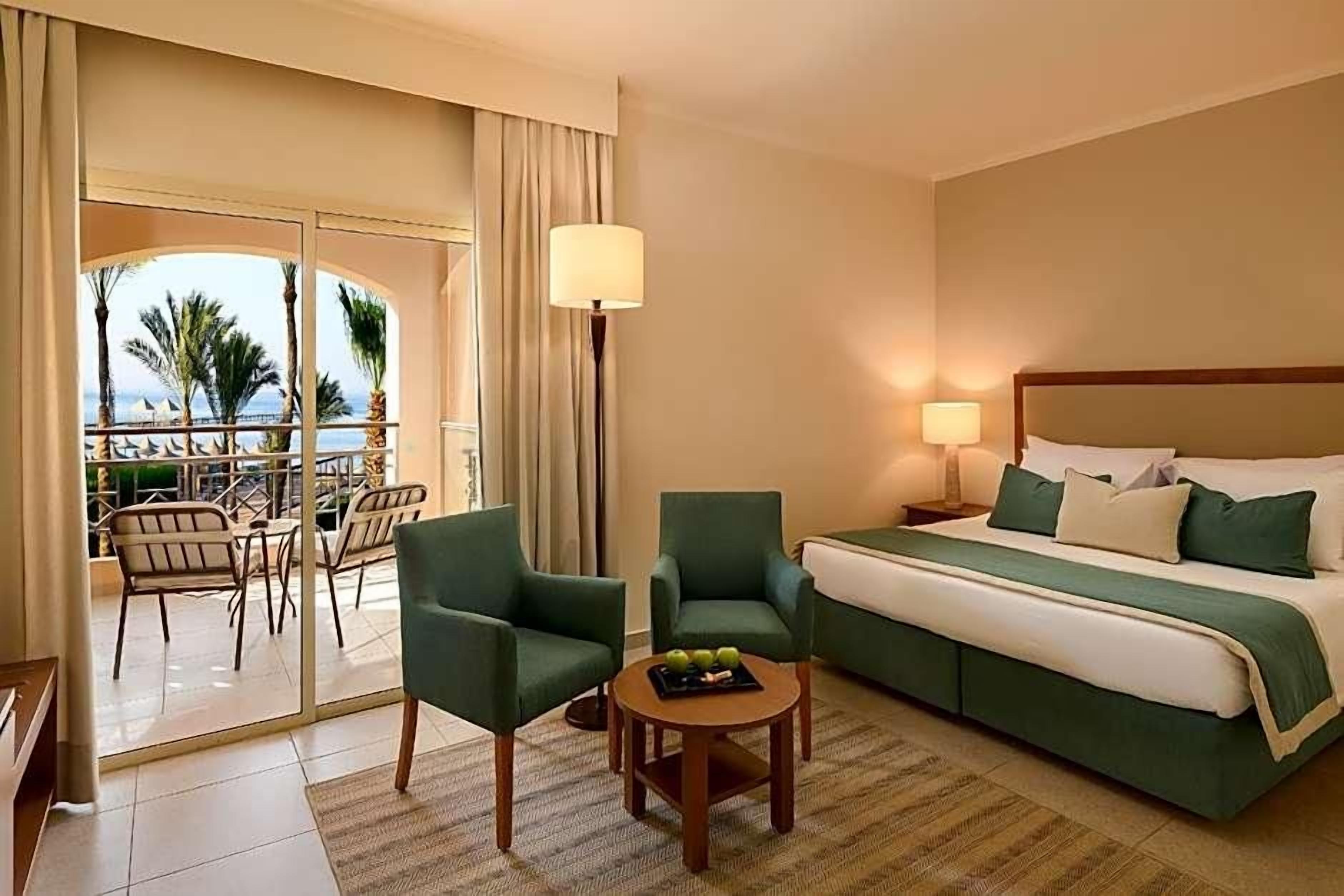 Une chambre d’hôtel avec un lit, deux fauteuils, une petite table avec des fruits, une lampe et une vue sur la mer par une porte vitrée.