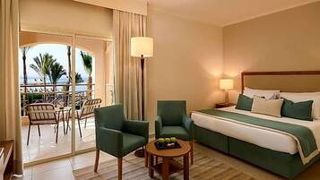 Une chambre d’hôtel avec un lit, deux fauteuils, une petite table avec des fruits, une lampe et une vue sur la mer par une porte vitrée.