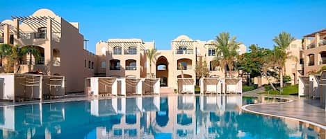 A pool area with lounge chairs and a clear blue pool, surrounded by buildings with arches and palm trees.