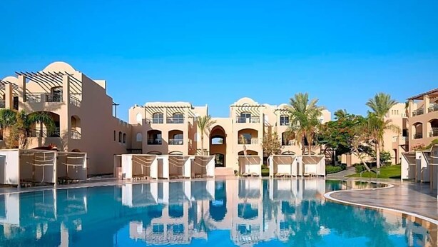 A pool area with lounge chairs and a clear blue pool, surrounded by buildings with arches and palm trees. - Magic World Sharm - Club By Jaz (Sharm El Sheikh)