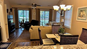 A spacious living area with a large sofa, a dining table with chairs, a ceiling fan, and a view of the outdoors through large windows. - Charming 2-bedroom condo in delightful Pawleys Island with AC, WiFi.   (Pawleys Island)