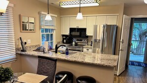 A modern kitchen with granite countertops, stainless steel appliances, and a dining area with a glass table and wicker chairs. - Charming 2-bedroom condo in delightful Pawleys Island with AC, WiFi.   (Pawleys Island)