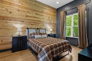 Quarto com parede de madeira, cama com colcha xadrez, duas mesas de cabeceira com abajures e uma janela com cortinas.
