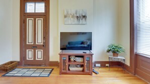 A living room with a wooden door, a flat-screen TV on a stand, a welcome sign, a potted plant, and a small table with a remote control. - STL Apartment: Close to Downtown! (St. Louis)