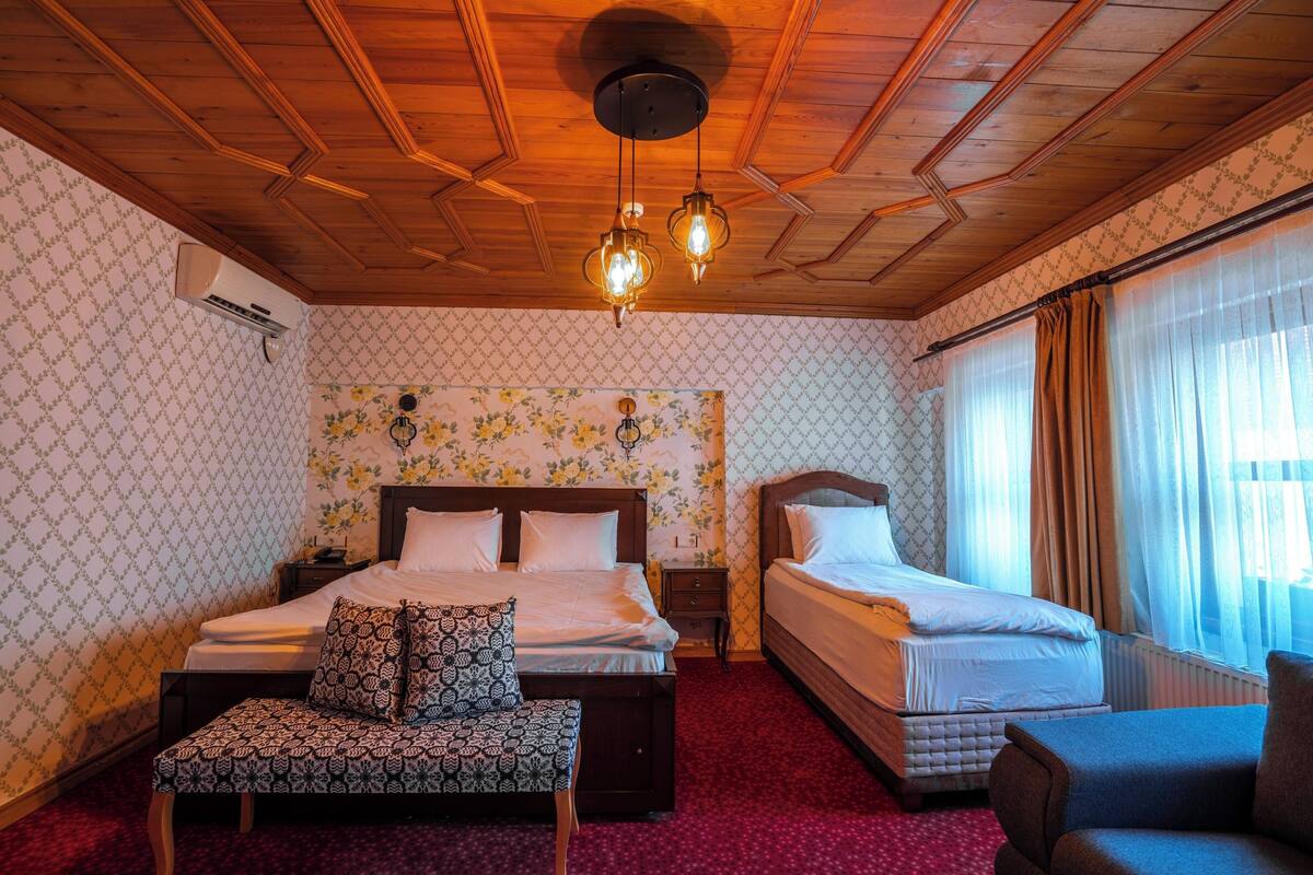 A hotel room with two beds, a bench, a wooden ceiling, and a floral wallpaper.