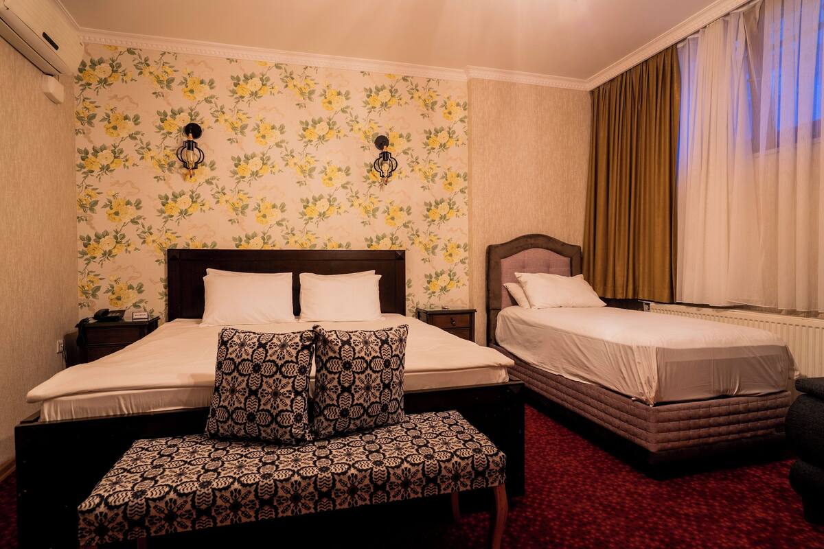A hotel room with two beds, a patterned bench, floral wallpaper, and a window with curtains.