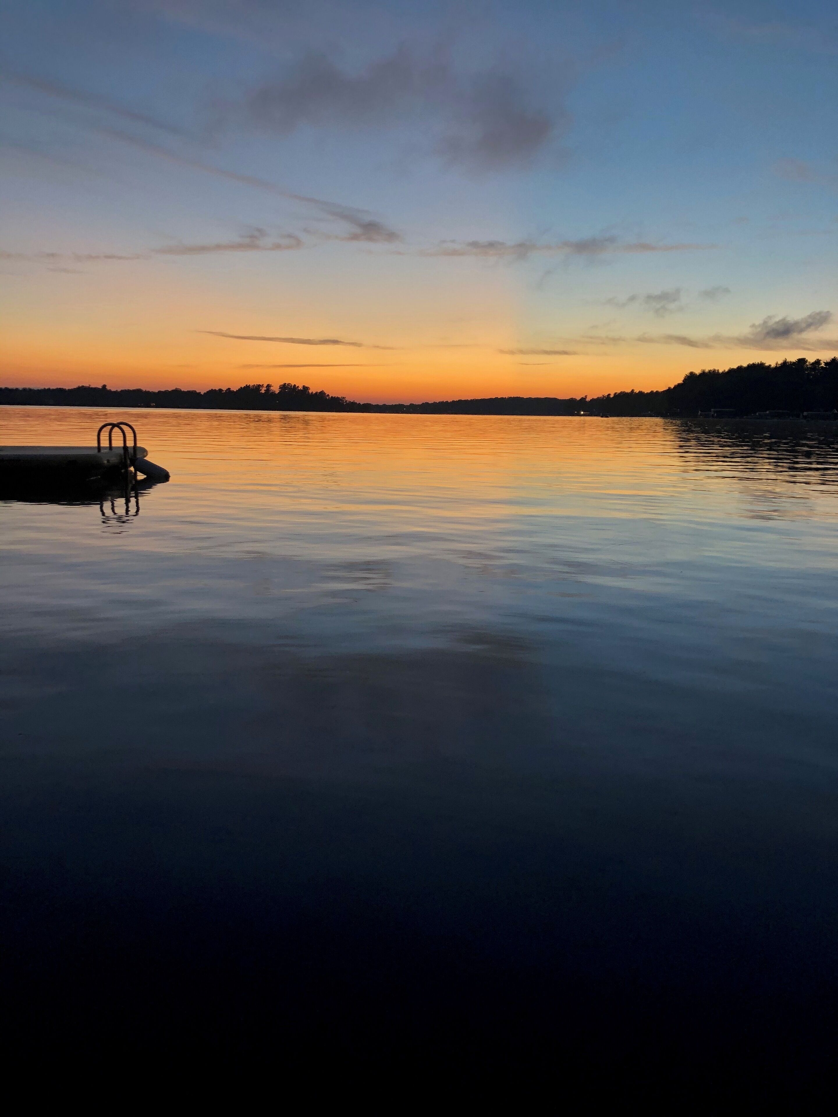 A calm lake at sunset with a dock and a row of trees in the background.
