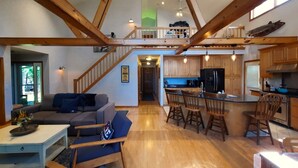 Amplia sala de estar con techo alto, vigas de madera, una escalera, una cocina con armarios de madera y un comedor con taburetes.