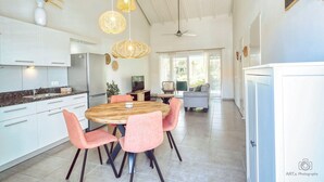 Eine moderne Küche mit weißen Schränken, einem Holztisch und rosa Stühlen. Hängende Korblichter und ein Deckenventilator sind vorhanden.