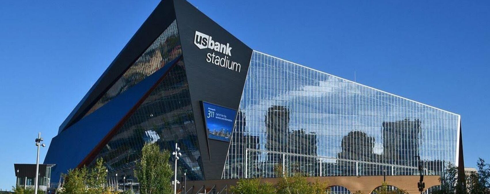 A modern building with a prominent 'US Bank Stadium' sign, featuring a unique angular design and large glass facade reflecting the surrounding cityscape.