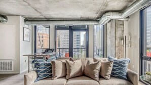 A modern living room with a beige sofa, large windows, and visible cityscape views. - CW  2BR 1B  Sky Lounge  Rooftop Pool  Gym  U (Minneapolis)