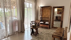 A spacious living room with a large window, a ceiling fan, a wooden sofa, a wooden table, and a woven coffee table. - M&m Villas Zanzibar (Kidoti)