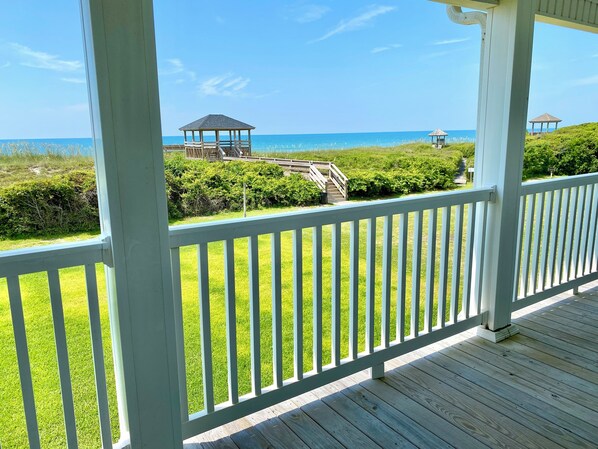 Une terrasse en bois avec une balustrade blanche donnant sur une plage où se trouvent deux kiosques et une pelouse.
