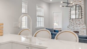A modern kitchen with white cabinetry, a white sink, and a white countertop. There are two wicker chairs with white cushions, a blue sofa, and a white wall with a brick pattern. - The Philip's Suite, newly renovated 3 Bed/3 EnSuite Bath, 1 Block to King (Charleston)