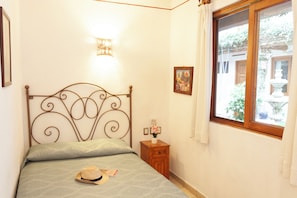 Un dormitorio con estructura metálica para la cama, una mesita de noche con lámpara, una ventana con cortinas y un pequeño cuadro en la pared.