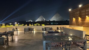 A rooftop terrace with tables and chairs, overlooking pyramids at night. - Pyramids Hotel (Giza)