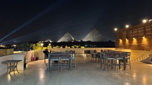 An outdoor seating area with tables and chairs, a wooden wall with lights, and two illuminated pyramids in the background. - Pyramids Hotel (Giza)
