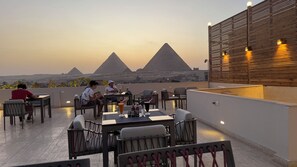 A rooftop terrace with tables and chairs, a wooden wall with lights, and a view of pyramids in the background. - Pyramids Hotel (Giza)