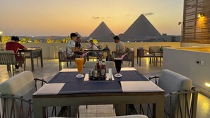 A rooftop dining area with tables and chairs, a view of the pyramids, and a table with condiments and drinks. - Pyramids Hotel (Giza)