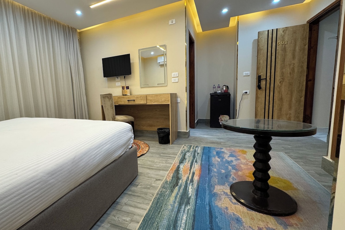 A hotel room with a bed, a desk with a chair, a TV, a small cabinet, and a round table.