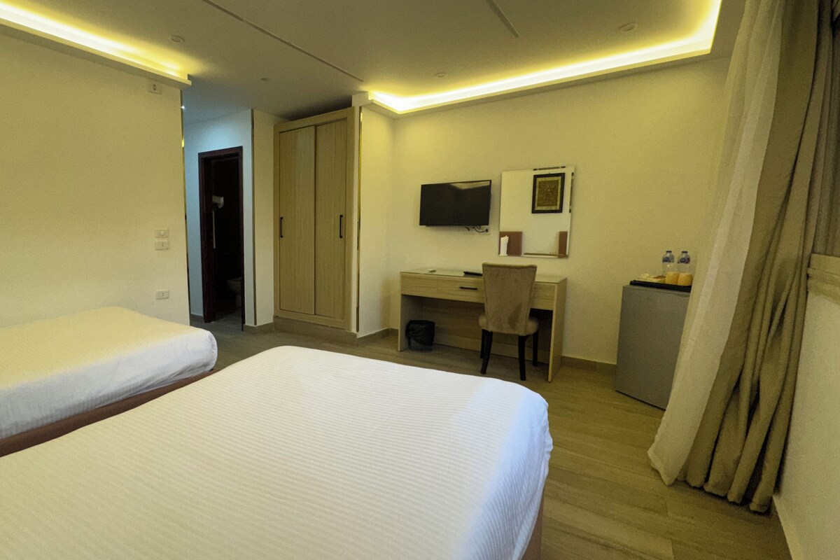A hotel room with two beds, a desk, a chair, a television, and a minibar.