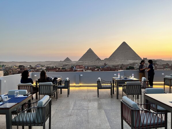 A rooftop dining area with tables and chairs, a view of the pyramids, and a cityscape in the background. - Pyramids Hotel (Giza)