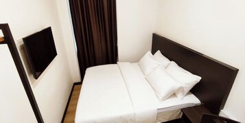 Uma cama bem arrumada com lençóis e almofadas brancas, uma cabeceira escura e uma TV de tela plana na parede.