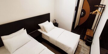 Habitación de hotel con dos camas individuales, una mesita de noche pequeña, un perchero y vista al interior.