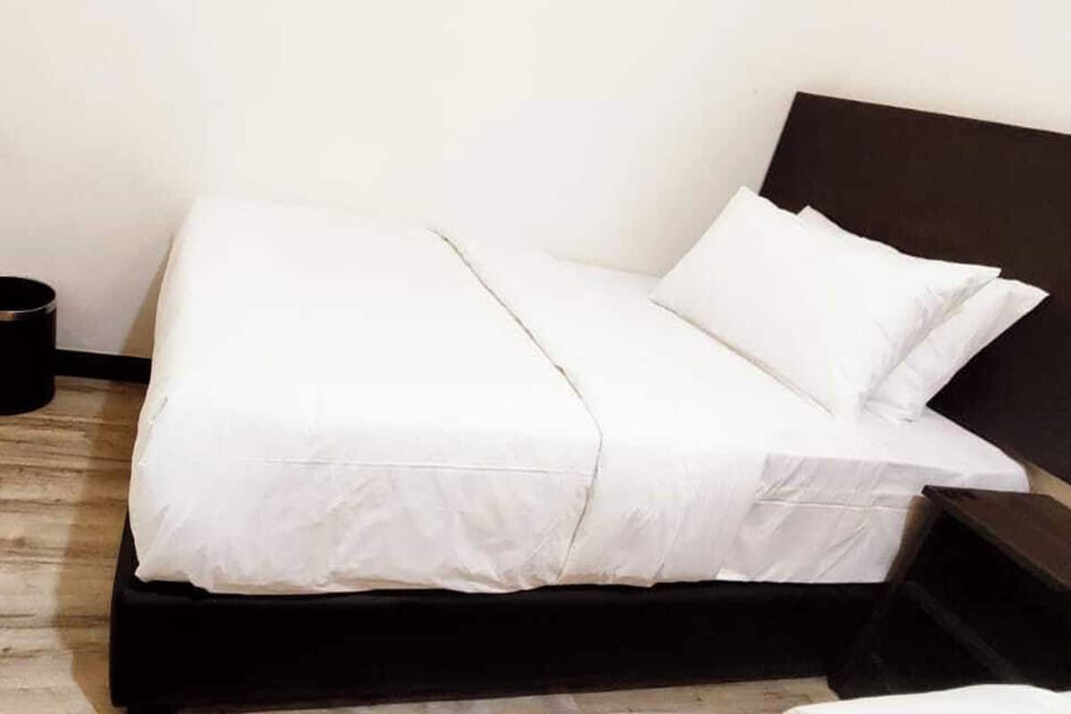 Uma cama de solteiro com roupa de cama branca num quarto com chão de madeira e cabeceira escura.