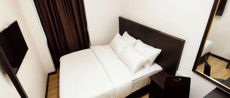A neatly made bed with white linens and pillows, a dark headboard, a bedside table with a black speaker, and a mirror on the wall.