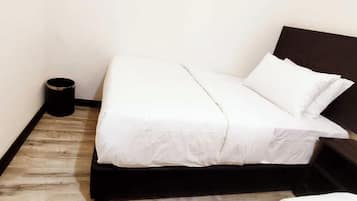 Un lit simple avec une literie blanche, dans une pièce dotée d’un plancher en bois et d’une tête de lit sombre.
