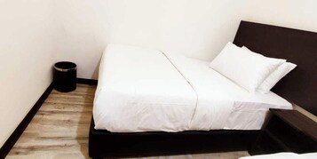 Cama de solteiro com roupa de cama branca num quarto com piso de madeira e cabeceira escura.