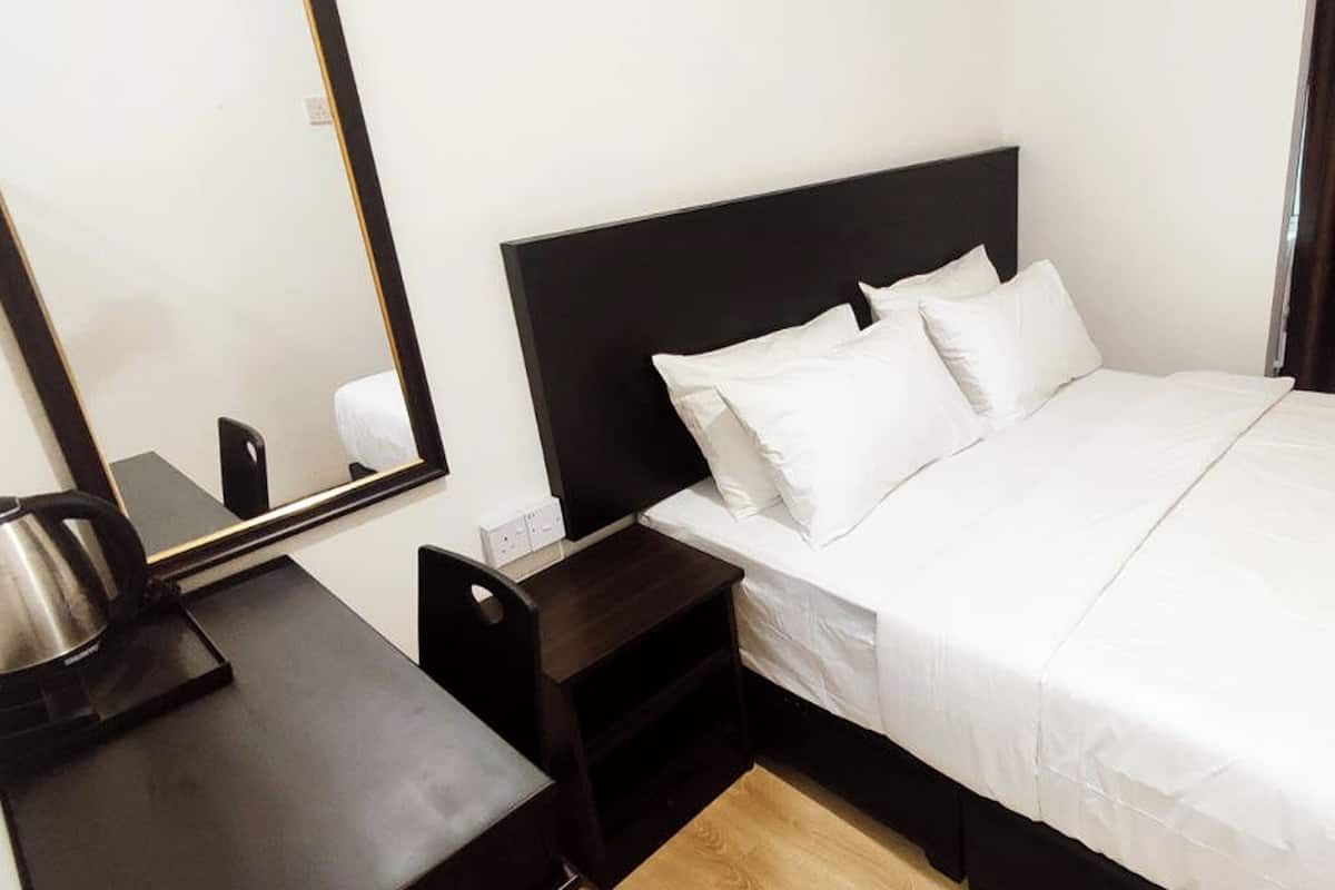 Quarto de hotel com uma cama, uma mesinha de cabeceira com chaleira, uma gavetinha e um espelho na parede.