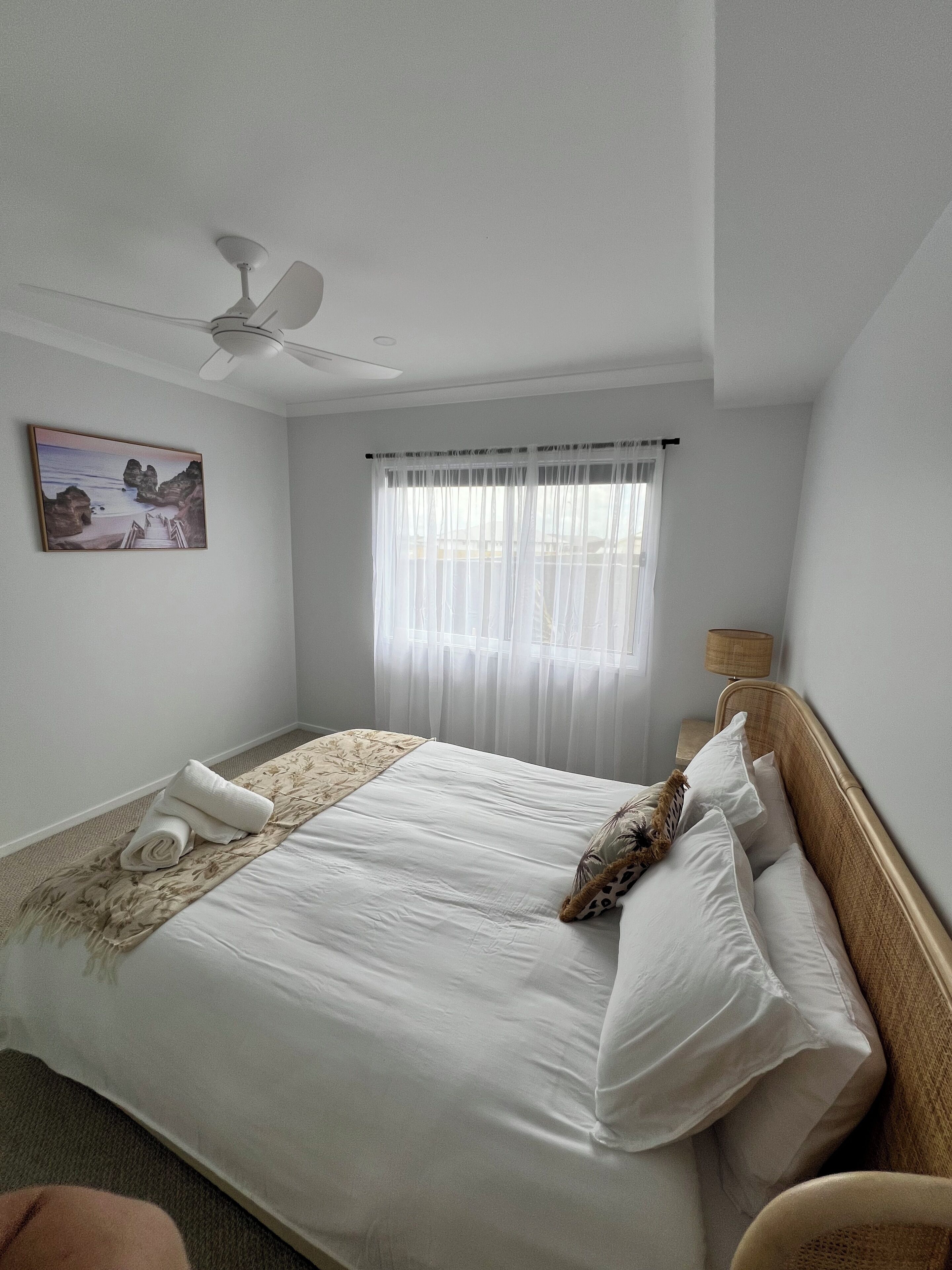 Een net opgemaakt bed met een bloemenbedekking, een kussen met een girafdesign, en een opgerolde handdoek ernaast. Er is een plafondventilator, een raam met dunne gordijnen en een schilderij van een strandtafereel aan de muur.
