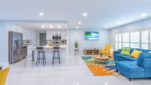 A modern kitchen with stainless steel appliances, a central island, and bar stools. There is a blue sofa with yellow cushions and a colorful rug in the living area. - Private Pool, Patio & BBQ Near Beaches & Casino (Margate)