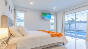 A modern bedroom with a large bed, a TV mounted on the wall, and a view of a swimming pool through the window. - Private Pool, Patio & BBQ Near Beaches & Casino (Margate)