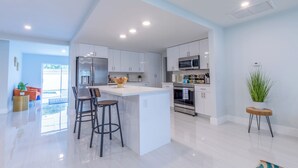 A modern kitchen with white cabinets, a central island with stools, stainless steel appliances, and a view to a bright room with orange furniture. - Private Pool, Patio & BBQ Near Beaches & Casino (Margate)