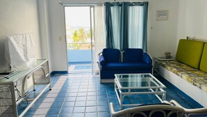 Una sala de estar con un sofá azul, un sofá verde, una mesa de centro blanca y un mueble blanco para el televisor. Hay una puerta corrediza de vidrio que da a un balcón con vista a palmeras.