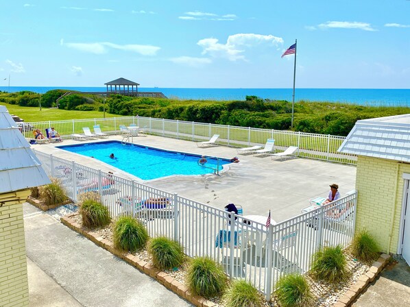 A swimming pool with a view of the ocean, surrounded by a white fence and lounge chairs.