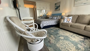 Une pièce chaleureuse avec un fauteuil en osier blanc, un canapé beige, un lit à motifs et un tapis bleu et beige.