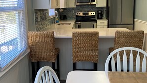 Une cuisine compacte avec des meubles blancs, des appareils en acier inoxydable et un coin repas doté de chaises en osier et d’une table blanche.