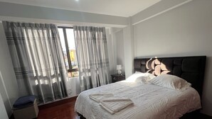 A neatly made bed with white linens, a wooden floor, and a window with grey curtains. - Skyline Suites (La Paz)
