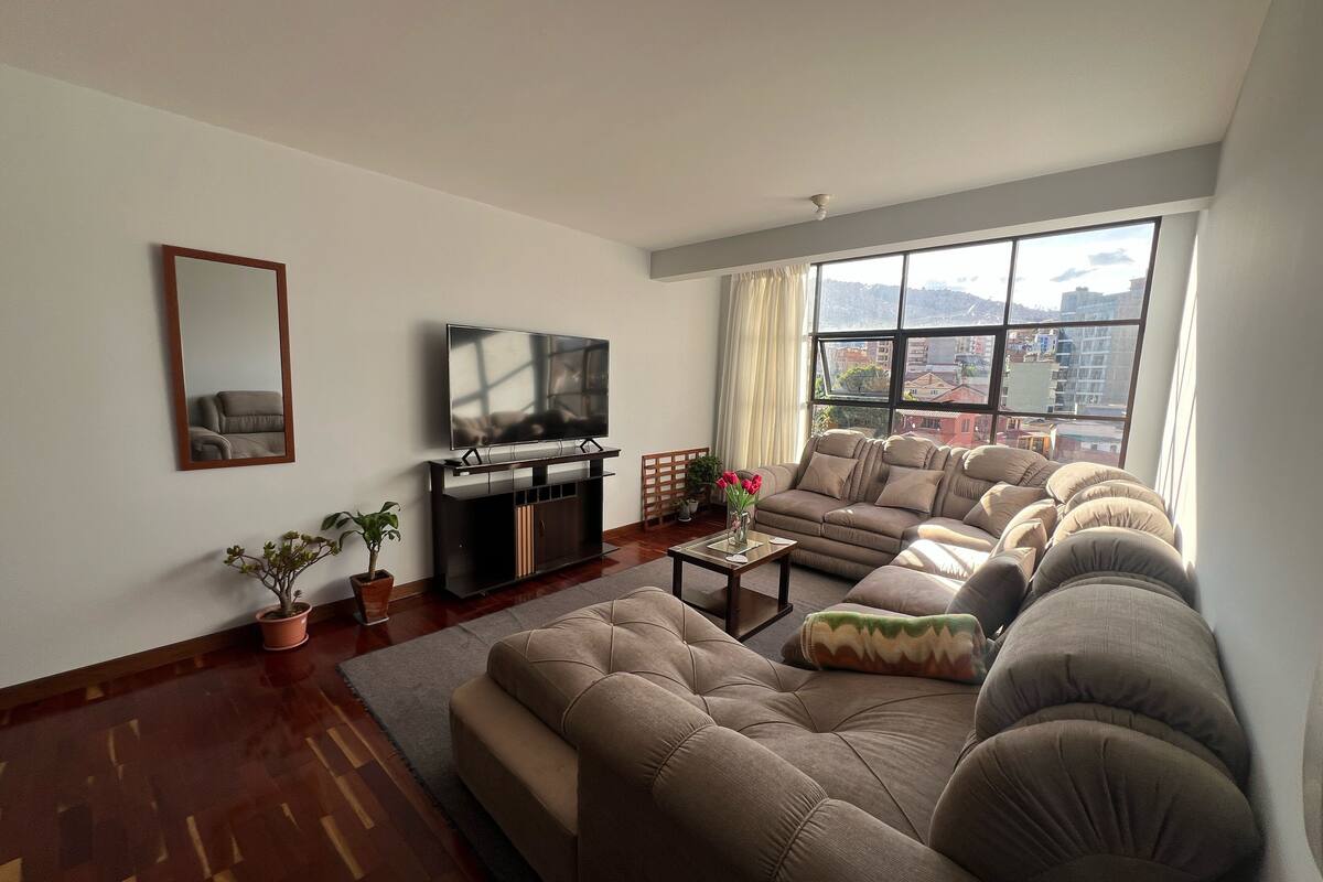 A living room with a sofa set, a TV on a stand, a mirror, and a window with a city view.