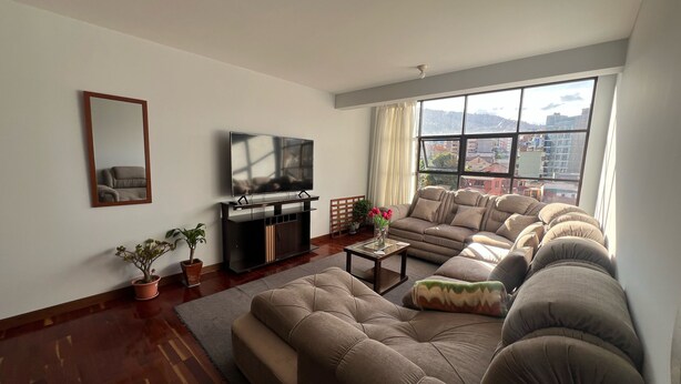 A living room with a sofa set, a TV on a stand, a mirror, and a window with a city view. - Skyline Suites (La Paz)