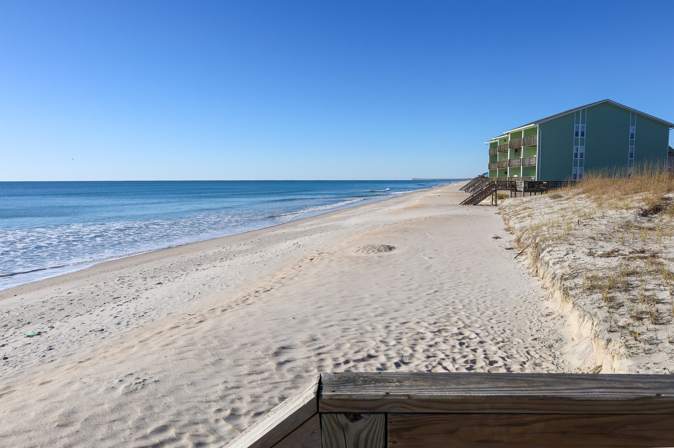 Una playa con un paseo de madera que lleva a un edificio verde con varios balcones.