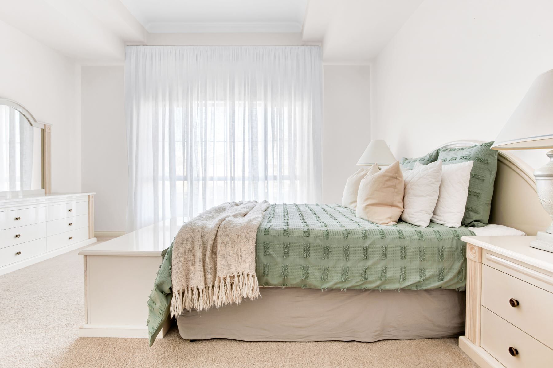 Une chambre à coucher comprenant un lit, une commode, une table de chevet et une fenêtre ornée de rideaux légers.