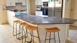 Ett modernt kök med vita skåp, en bänkskiva i marmor, ett kylskåp i rostfritt stål och en inbyggd ugn. Vid köksön finns tre trästolar.