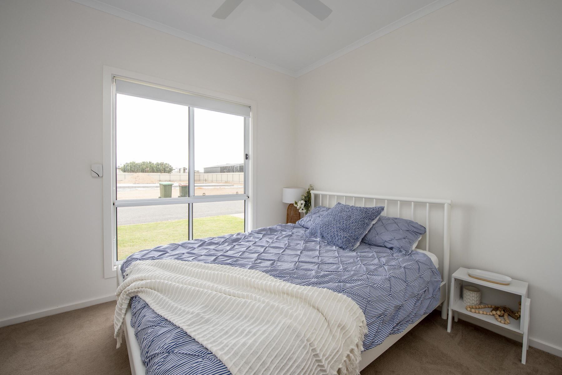 A bedroom with a large window, a bed with blue and white bedding, a white bedside table with a lamp, and a small shelf with decorative items.