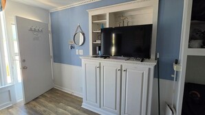 A room with a white cabinet, a flat-screen TV, a mirror, and a shelf with items.